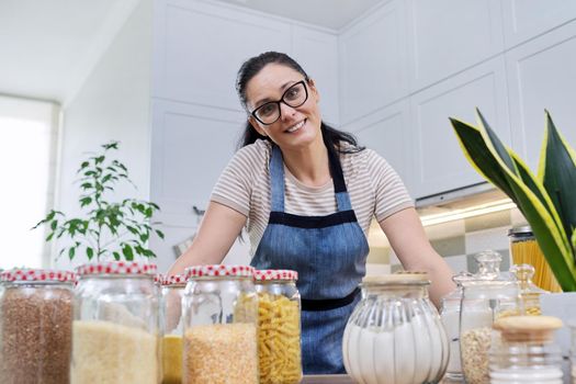 Storing food in kitchen, woman housewife in an apron with jars and containers of cereals, food, pasta, talking and looking at camera. Female blogging about organizing space in kitchen