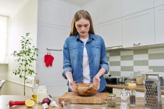 Young woman prepares chicken at home, marinates meat with spices in glass bowl on table, kitchen interior background, cook at home