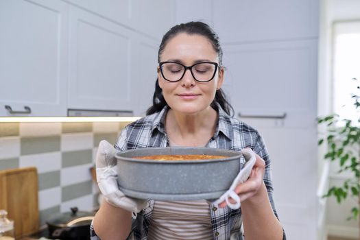 Portrait of smiling woman in kitchen mittens holding hot freshly baked apple pie, happy housewife with glasses enjoying the smell of homemade baked goods
