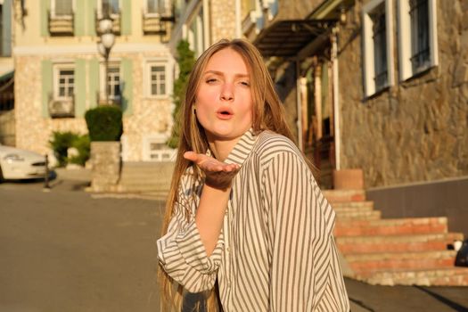 Young beautiful blonde sending a passionate kiss to the camera, sunset summer city street background. Body language, gestures, beauty, love, people, emotions concept