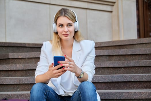 Young beautiful woman in headphones with smartphone resting sitting on steps in city, listening music watching video. Happiness, relaxation, lifestyle, youth and technology