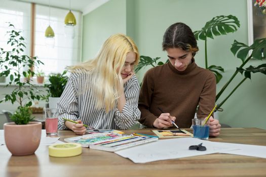 Teenagers painting with watercolors. Couple of guy and girl sitting at table with paints, brushes, drawings. Happy teens talking laughing creative. Creativity, hobby, leisure concept