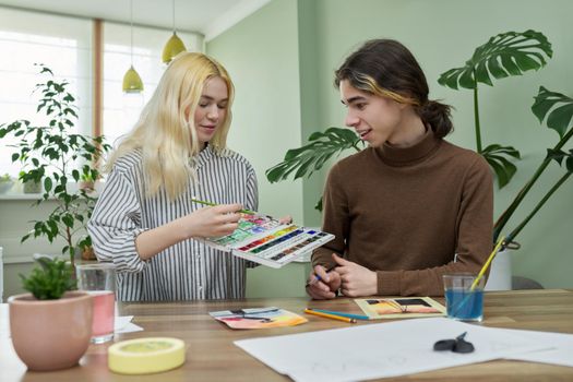 Teenagers painting with watercolors. Couple of guy and girl sitting at table with paints, brushes, drawings. Happy teens talking laughing creative. Creativity, hobby, leisure concept