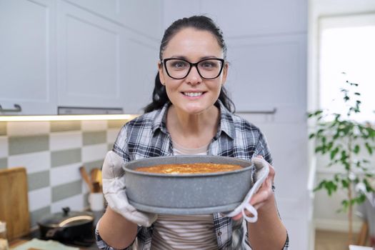 Portrait of smiling woman in kitchen mittens holding hot freshly baked apple pie, happy housewife with glasses enjoying the smell of homemade baked goods