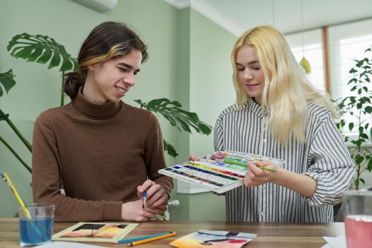 Teenagers painting with watercolors. Couple of guy and girl sitting at table with paints, brushes, drawings. Happy teens talking laughing creative. Creativity, hobby, leisure concept