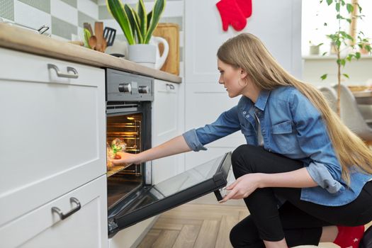 Cooking at home, roasting meat in oven, young woman putting marinated chicken in baking bag in oven, kitchen interior background