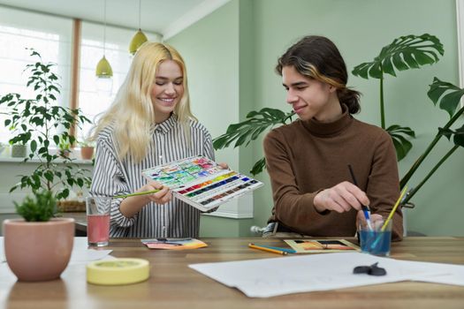 Teenagers painting with watercolors. Couple of guy and girl sitting at table with paints, brushes, drawings. Happy teens talking laughing creative. Creativity, hobby, leisure concept