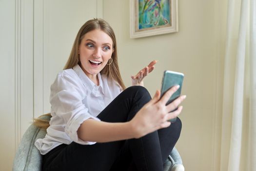 Young beautiful blonde sitting at home in chair, laughing talking using video communication on smartphone. Leisure, woman relaxing looking at phone screen