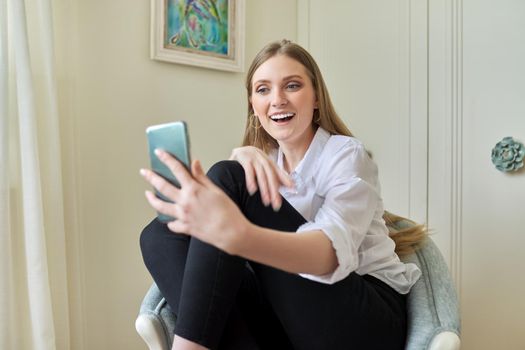 Young beautiful blonde sitting at home in chair, laughing talking using video communication on smartphone. Leisure, woman relaxing looking at phone screen