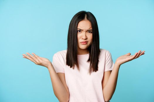 Confused and annoyed asian girl shrugging shoulders, furrow eyebrows and spread hands puzzled, cant understand smth, blue background.