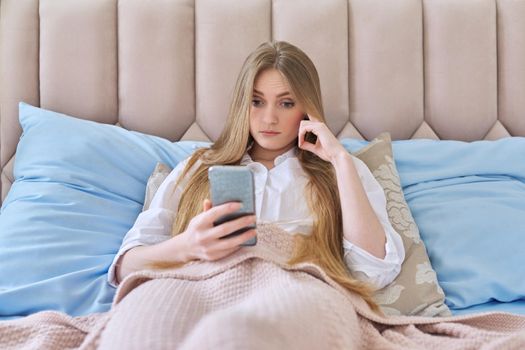 Serious surprised young woman with smartphone resting lying at home in bed under a blanket reading, focused