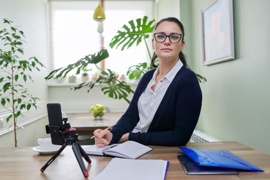 Business woman looking in smartphone webcam on tripod at home. Female talking online recording video blog, consulting finance, mentoring teaching training remotely, helping psychologist, distance work
