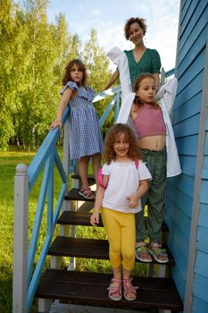 Cheerful woman with adorable girls standing on stairs outside blue cottage on sunny summer day in countryside