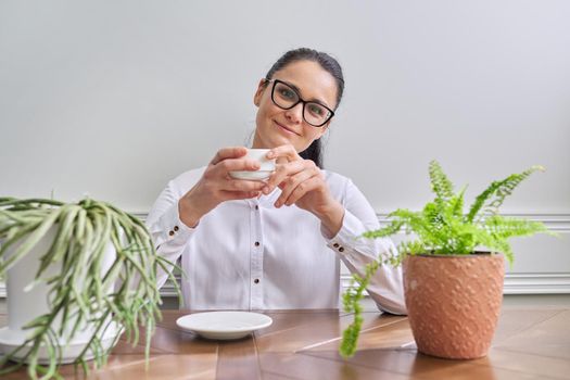 Talking positive business woman looking at camera. Middle-aged female wearing white shirt glasses sitting at table with cup of coffee, gesturing making video consultation, recording video vlog