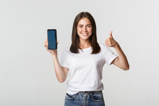Satisfied smiling attractive woman showing smartphone screen and thumbs-up.