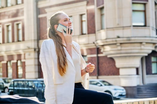 Beautiful young business woman talking on cell phone in city. Smiling successful happy blonde woman, building background in sunset light