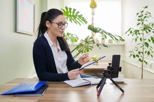 Business woman looking in smartphone webcam on tripod at home. Female talking online recording video blog, consulting finance, mentoring teaching training remotely, helping psychologist, distance work