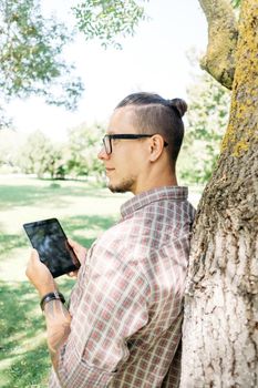 Young man in glasses and plaid shirt standing near a tree with digital tablet in summer park.