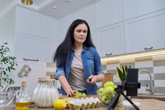 Woman cooking apple pie at home in the kitchen, talking online using a video call on a smartphone.