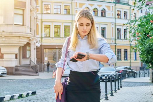 Young business woman in city looking at her wristwatch. Confident serious female with smartphone in her hands, with laptop bag, sunset in the city, urban style background