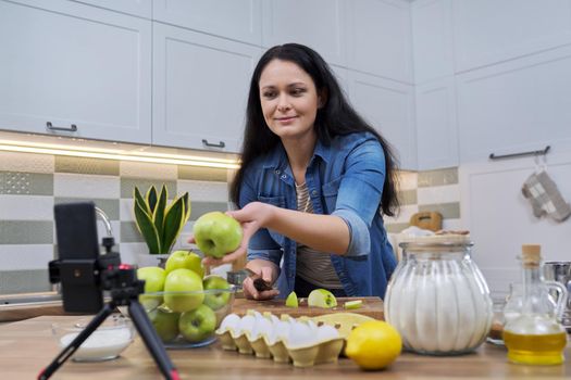 Woman cooking apple pie at home in the kitchen, talking online using a video call on a smartphone, showing green apples on webcam