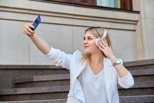 Young beautiful woman in headphones with smartphone resting sitting on steps in city, listening music watching video. Happiness, relaxation, lifestyle, youth and technology
