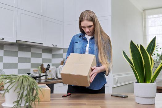 Young happy woman unpacking cardboard boxes, unboxing expected postal parcel with online shopping, at home in kitchen