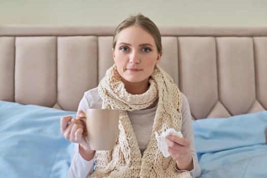 Sick young woman sitting at home in bed with hot cup of tea and handkerchief. Seasonal colds, cough, runny nose, viral infections, home treatment