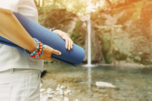 Unrecognizable woman with yoga mat standing in front of waterfall on sunny summer day outdoor.