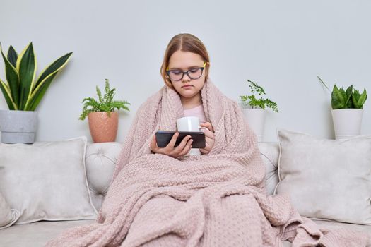 Cold autumn winter season at home, teenage girl under warm knitted blanket sitting on sofa with mug of hot tea and smartphone