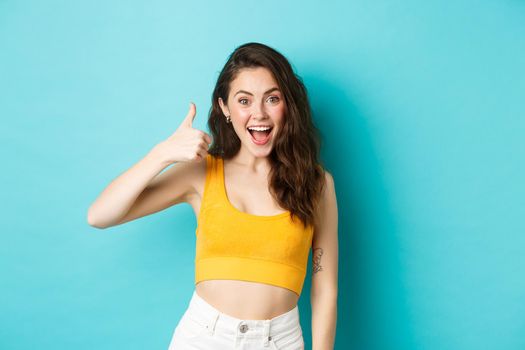 Summer holidays and emotions concept. Cheerful young woman saying yes, praising good choice, nice work, showing thumbs up and looking excited, standing over blue background.