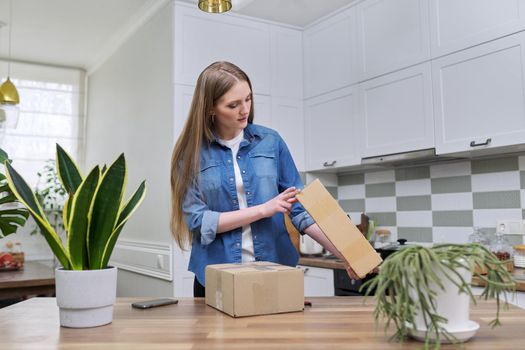 Young happy woman unpacking cardboard boxes, unboxing expected postal parcel with online shopping, at home in kitchen