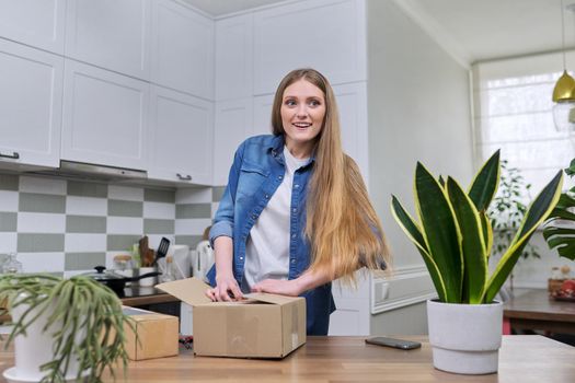 Young happy woman unpacking cardboard boxes, unboxing expected postal parcel with online shopping, at home in kitchen