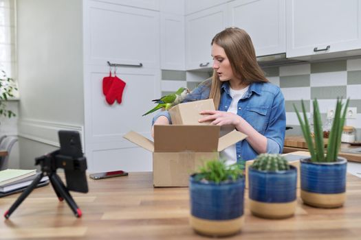 Woman recording video on smartphone moment of unpacking cardboard box. Showing purchases ordered on Internet, product quality reviews with followers, blog content, feedback. Pet parrot helping unpack