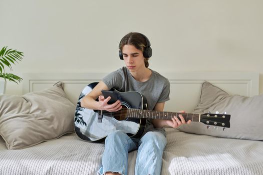 Male teenager in headphones sitting at home on bed with acoustic guitar and smartphone. Hipster guy using smartphone to learn music, study songs, record his music. Creativity, hobby, teens concept