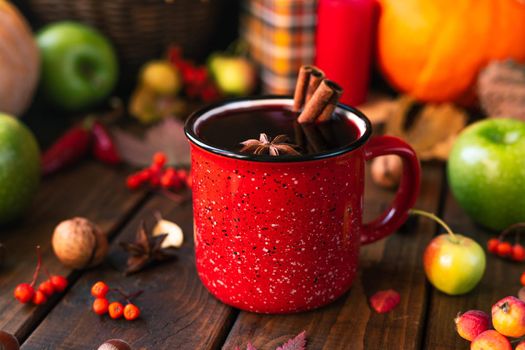 red mug with mulled wine. Cinnamon sticks stick out of the cup and a star of star anise floats. Fruits and spices are all around on a table