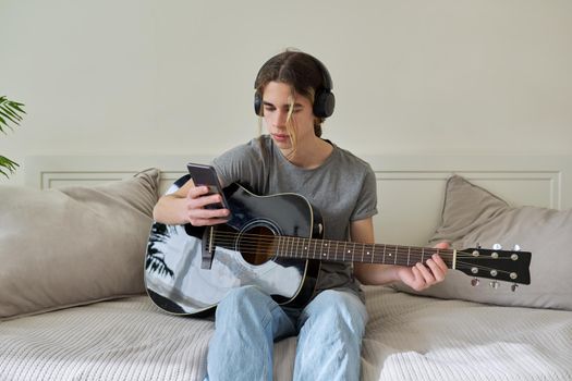 Male teenager in headphones sitting at home on bed with acoustic guitar and smartphone. Hipster guy using smartphone to learn music, study songs, record his music. Creativity, hobby, teens concept