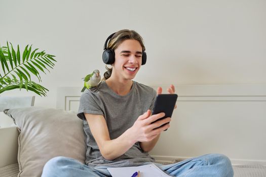 Emotional talking male teenager in headphones looking at smartphone screen using video call. Bird pet green quaker parrot on the owners shoulder, home room sofa bed background