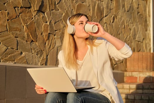 Young beautiful female in headphones with laptop drinking coffee in paper cup. Outdoor portrait of smiling woman sitting on steps in city