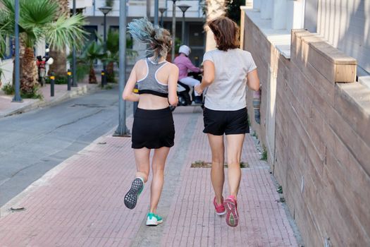 Running mother and teenage daughter, back view, along the street of a sunny summer city. Active healthy lifestyle, family and sports