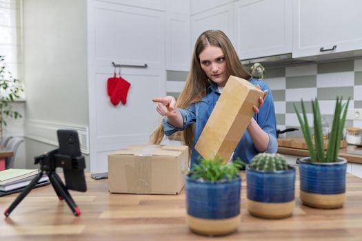 Woman recording video on smartphone moment of unpacking cardboard box. Showing purchases ordered on Internet, product quality reviews with followers, blog content, feedback. Pet parrot helping unpack