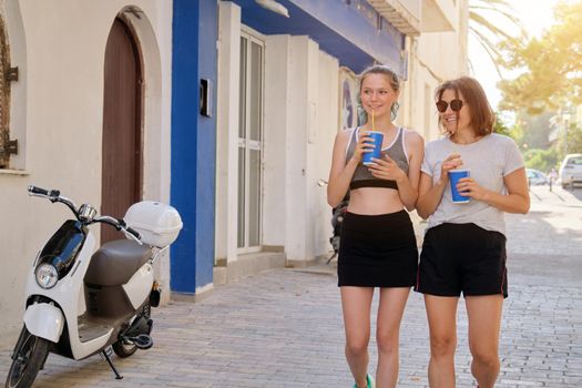 Mother and teenage daughter together in sportswear walking along the city street after fitness, jogging, talking laughing drinking drinks from paper cup