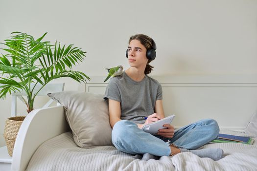 Male teenager with pet green parrot quaker on his shoulder, guy student sitting in headphones with textbooks notebooks books at home on bed