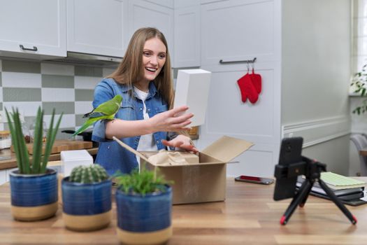 Woman recording video on smartphone moment of unpacking cardboard box. Showing purchases ordered on Internet, product quality reviews with followers, blog content, feedback. Pet parrot helping unpack