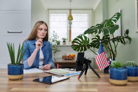 Young woman university student studying at home online remotely, e-learning. Talking female looking at smartphone screen, USA flag background, distance concept