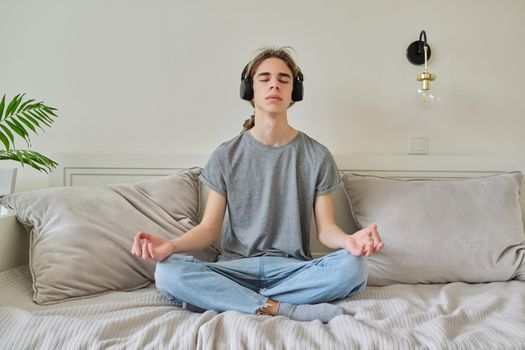 Male teenager sitting in lotus position at home on bed relaxing meditating with closed eyes wearing headphones