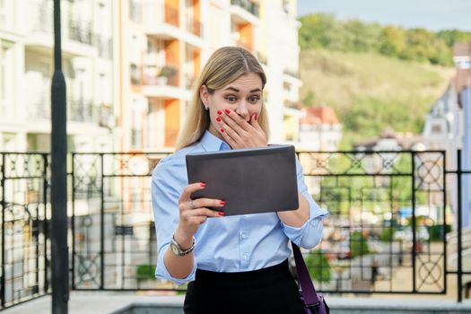 Surprised young business woman looking emotionally into digital tablet screen, on city street