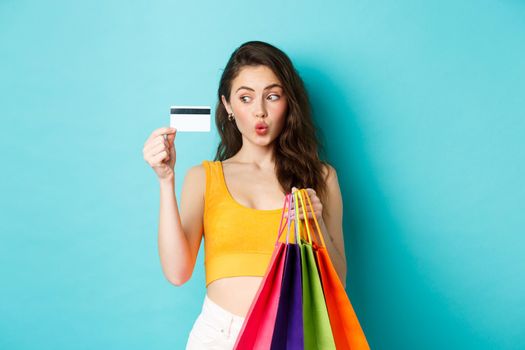 Image of happy woman shopaholic showing her plastic credit card, holding shopping bags, wearing summer clothes, standing against blue background.