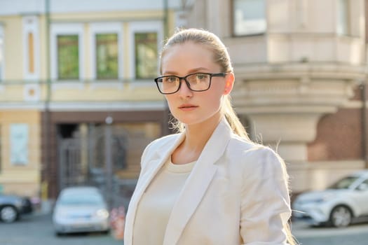 Portrait of young beautiful successful business woman looking at camera, smiling blonde in glasses in white jacket on street of sunny city