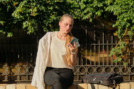 Outdoor sad serious young woman talking on smartphone, business female with laptop bag sitting in city, sunset light background